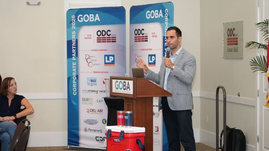 A man in a gray jacket speaks at the GOBA podium in front of sponsor banners.