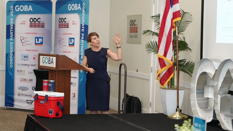 A woman gestures while presenting beside the GOBA podium and sponsor banners.