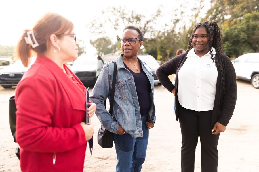Three guests talking outdoors at the Apopka home dedication, with a woman in denim in focus.