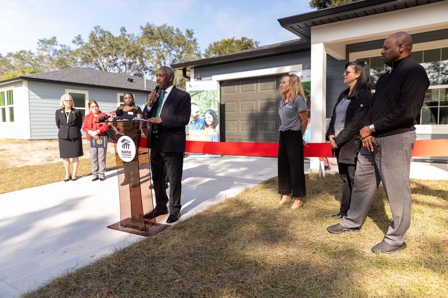 A speaker addresses the home-dedication gathering from a Habitat lectern beside a red ribbon.