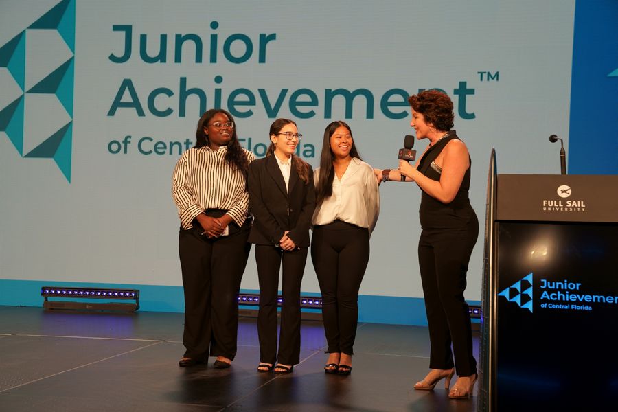A speaker addressing three young participants on stage.