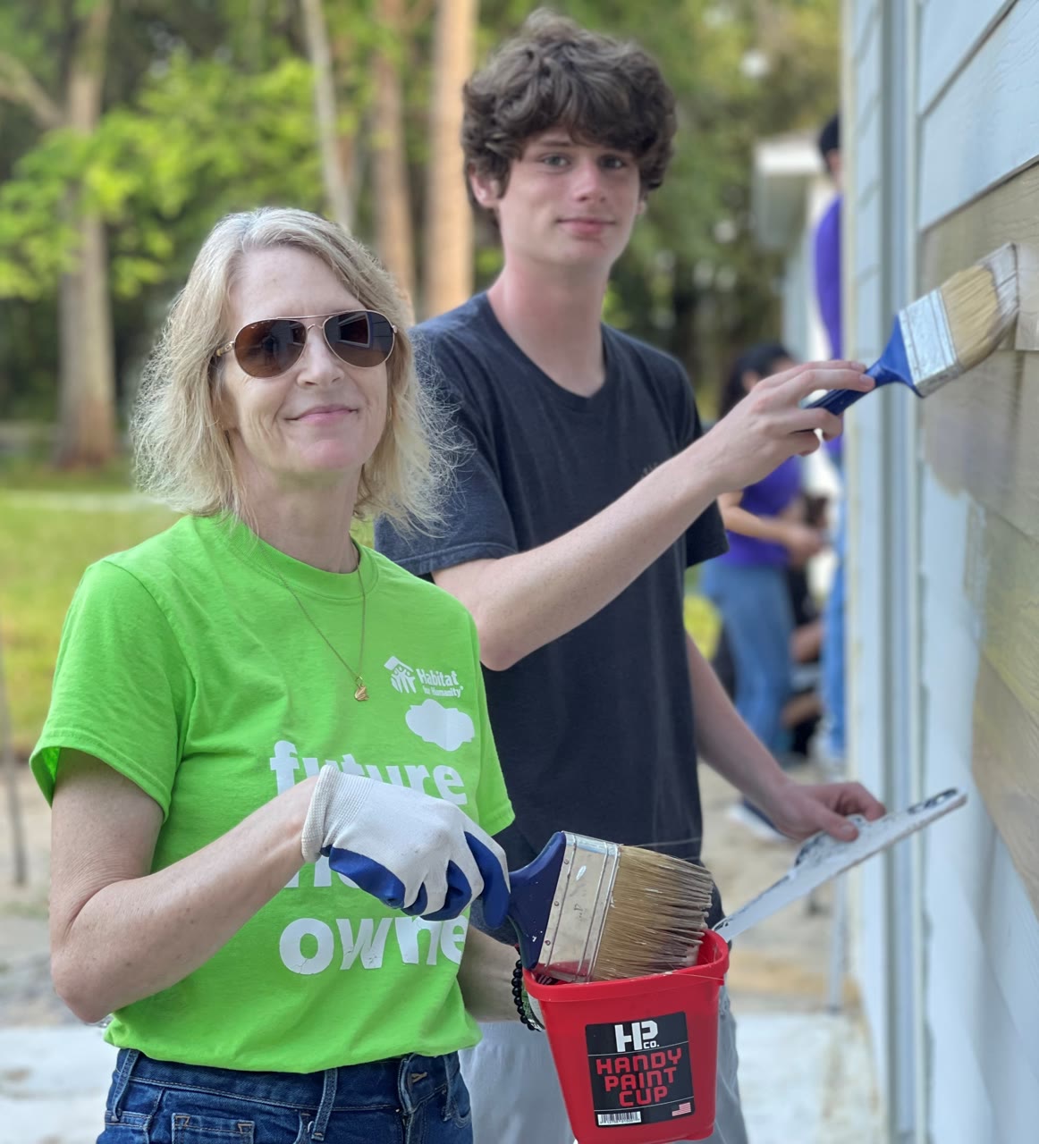 Two volunteers with paintbrushes beside a house; one wears a Habitat for Humanity shirt.